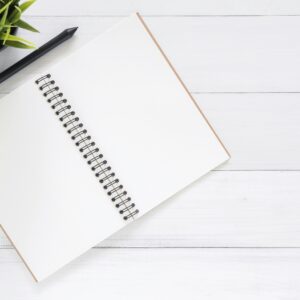 Open spiral notebook on white desk with black pen and green plant. Ideal for planning.